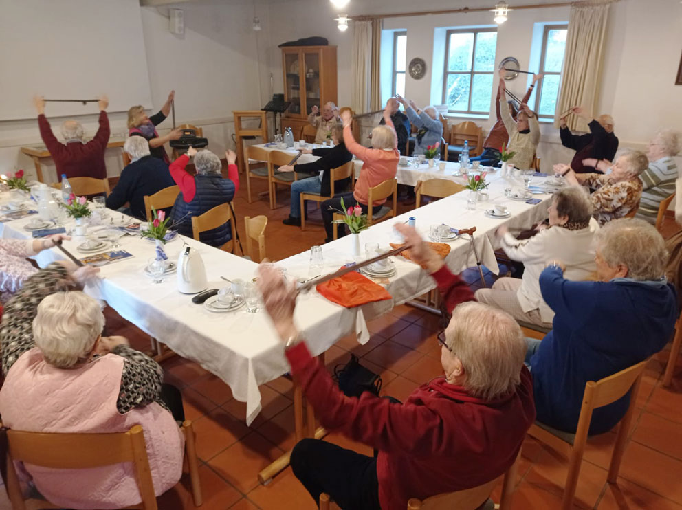 Senioren an Tischen sitzend, Übungen ausführend.